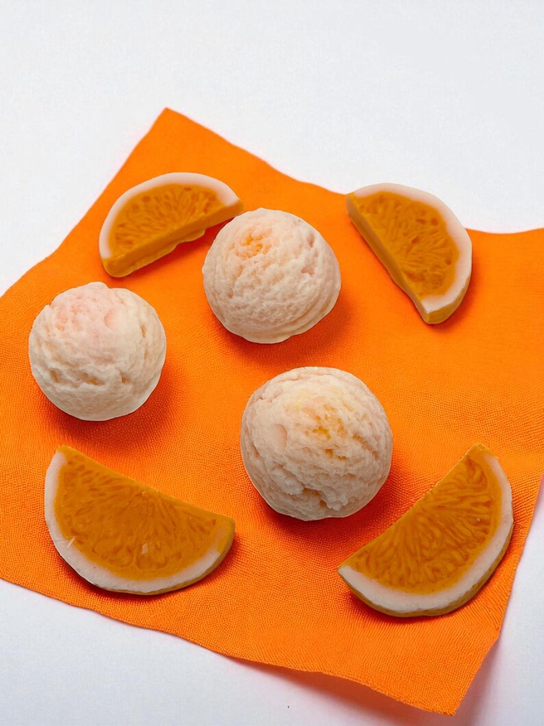 Op de afbeelding: Een compositie van sinaasappelschijfjes en ronde, lichtgekleurde bollen op een oranje doek. De schijfjes hebben een witte schil en een oranje binnenkant. De bollen hebben een textuur, mogelijk een voedingsmiddel.
