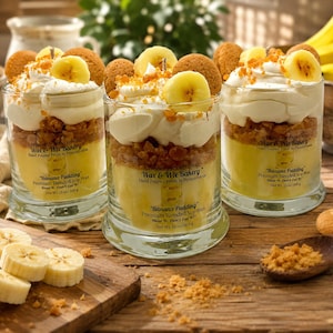 May include: Three glass jar candles labeled Wax & Wix Bakery Banana Pudding. Each candle features layers of yellow wax, brown crumbles, white whipped wax, banana slices, and round cookies. The labels state Net Wt. 13 oz/369 g.