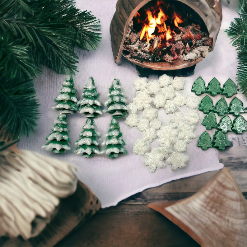 May include: A festive arrangement featuring miniature green Christmas trees with white accents, snowflake-shaped decorations, and additional green tree-shaped ornaments. The scene is set with a backdrop of a cozy fireplace and evergreen branches.