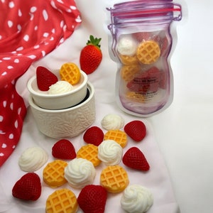 May include: A white ceramic wax warmer with a red strawberry and a waffle-shaped wax melt inside. A clear jar with a purple lid contains a variety of waffle and strawberry-shaped wax melts. The text on the jar reads "Strawberry Shortcake" and "Wax & Wick Bakery".
