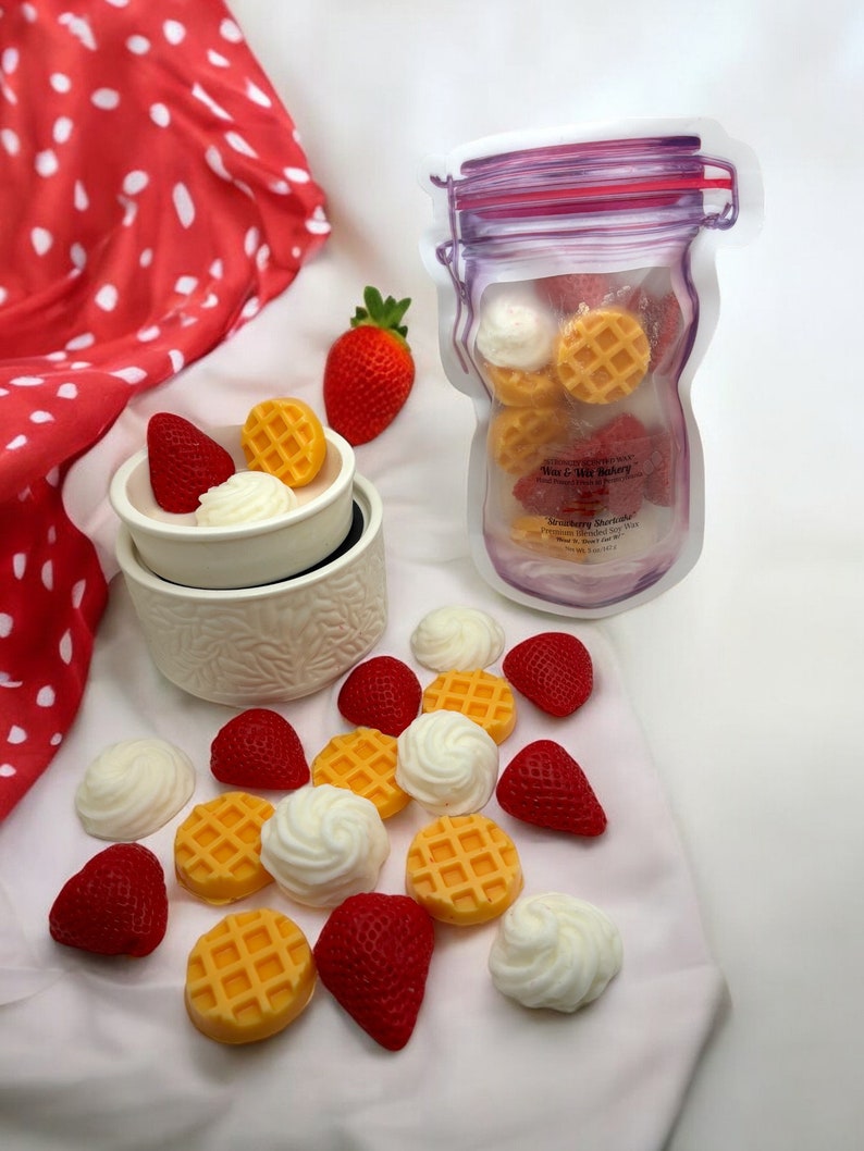 May include: A white ceramic wax warmer with a red strawberry and a waffle-shaped wax melt inside. A clear jar with a purple lid contains a variety of waffle and strawberry-shaped wax melts. The text on the jar reads "Strawberry Shortcake" and "Wax & Wick Bakery".