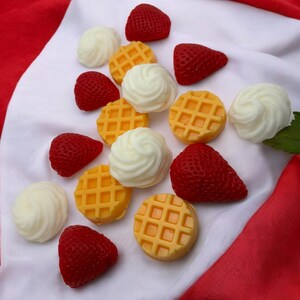 May include: Assortment of colorful, handmade soap bars shaped like strawberries and waffles. The soaps are arranged on a white and red fabric background.