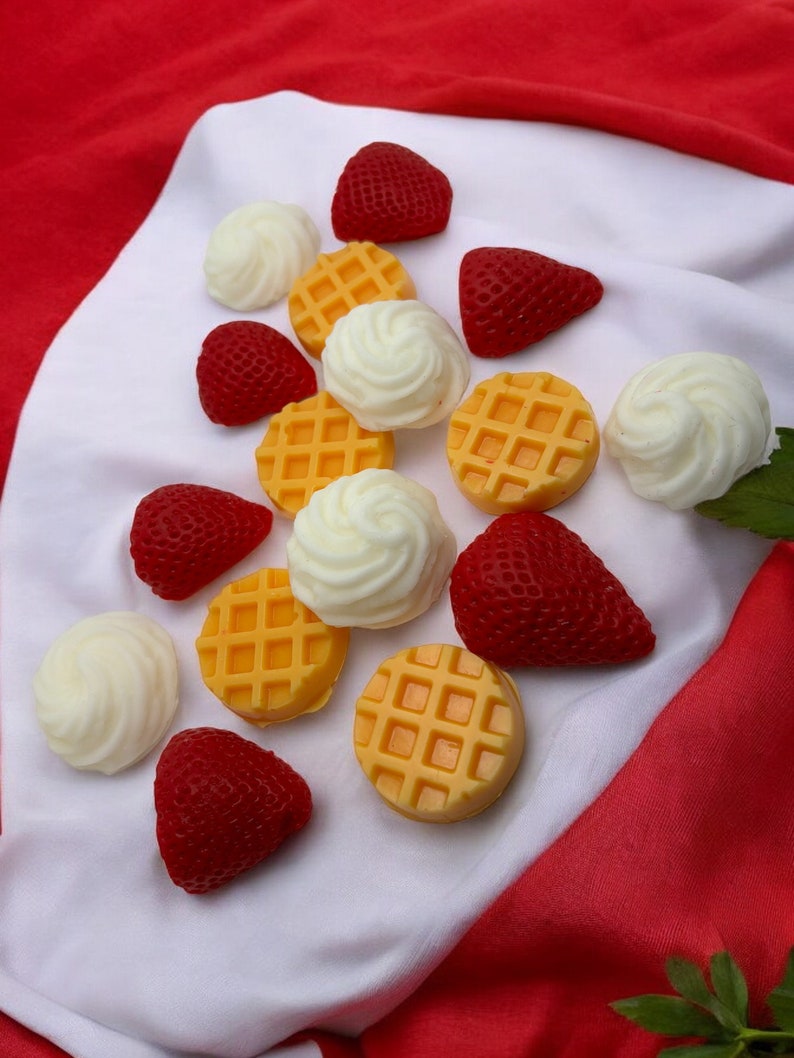 May include: Assortment of colorful, handmade soap bars shaped like strawberries and waffles. The soaps are arranged on a white and red fabric background.