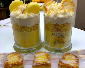 May include: Two glass jars filled with a lemon pound cake scented candle. The candle is layered with white frosting, yellow cake crumbs, and a yellow candle. The jars are topped with a lemon slice and a white frosting swirl. The jars are sitting on a white and brown striped fabric. The label on the jar reads "STRONGLY SCENTED WAX Wax & Wix Bakery Hand Poured Fresh in Pennsylvania "lemon Pound Cake" Premium Blended Soy Wax About 9 oz. Don't Eat Wax Net Wt. 1.1 oz/30g"