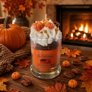 May include: A clear glass candle jar filled with brown wax, topped with white whipped wax and small orange pumpkin-shaped wax pieces. The orange label reads Wax & Wix Bakery, Pumpkin Pecan Waffles, and 12 oz. A single white wick is centered on top.