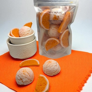 Op de afbeelding: Een doorzichtige zak gevuld met oranje-vormige wassmelt en sinaasappelschijfjes, met de tekst "Orange Sherbet Creamlete". Verschillende wassmelt en sinaasappelschijfjes zijn gerangschikt op een oranje doek.