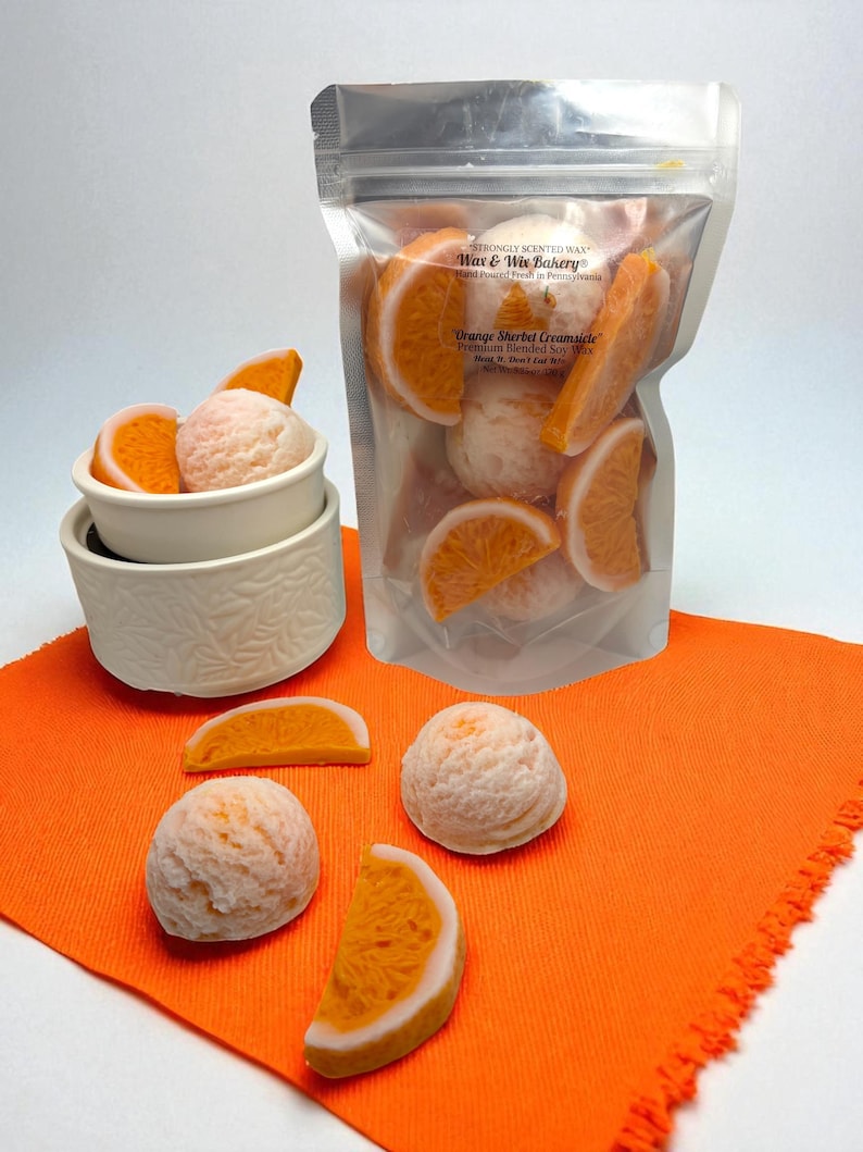 Op de afbeelding: Een doorzichtige zak gevuld met oranje-vormige wassmelt en sinaasappelschijfjes, met de tekst "Orange Sherbet Creamlete". Verschillende wassmelt en sinaasappelschijfjes zijn gerangschikt op een oranje doek.