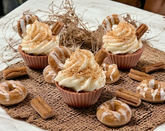 May include: A close-up of several decorative cupcakes with white frosting, sprinkles, and donut accents. The cupcakes are in pink holders and are surrounded by mini donuts and rectangular treats. The image is set on a brown woven surface.