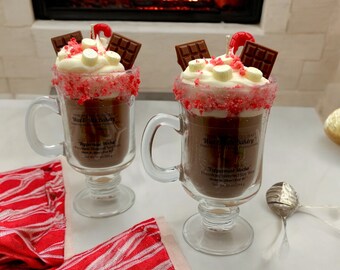 May include: Two clear glass mugs filled with a dark liquid, topped with whipped cream, marshmallows, chocolate pieces, and a red cherry. The rims are coated with red sprinkles. The mugs sit on a white surface with a red and white striped cloth in the foreground. The background features a fireplace.