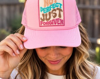 May include: A bright pink trucker hat featuring a rectangular patch on the front panel. The patch displays the text "NOT PERFECT JUST FORGIVEN" in layered, multi-colored embroidered lettering with tan, teal, brown, and pink hues.