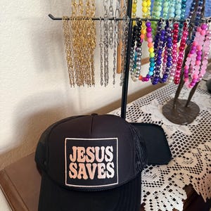 May include: A black trucker hat with a mesh back and a square patch that reads "JESUS SAVES" in a retro font. The hat sits on a wooden surface, with a jewelry display stand holding various necklaces in the background.