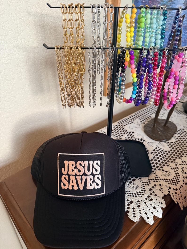 May include: A black trucker hat with a mesh back and a square patch that reads "JESUS SAVES" in a retro font. The hat sits on a wooden surface, with a jewelry display stand holding various necklaces in the background.