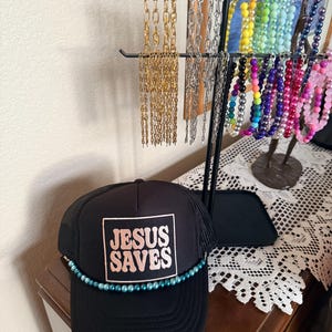 May include: Black trucker hat with the text "JESUS SAVES" in a square, with a teal bead trim. A jewelry display stand holds various necklaces in gold, silver, and colorful beads. The hat is on a wooden surface.