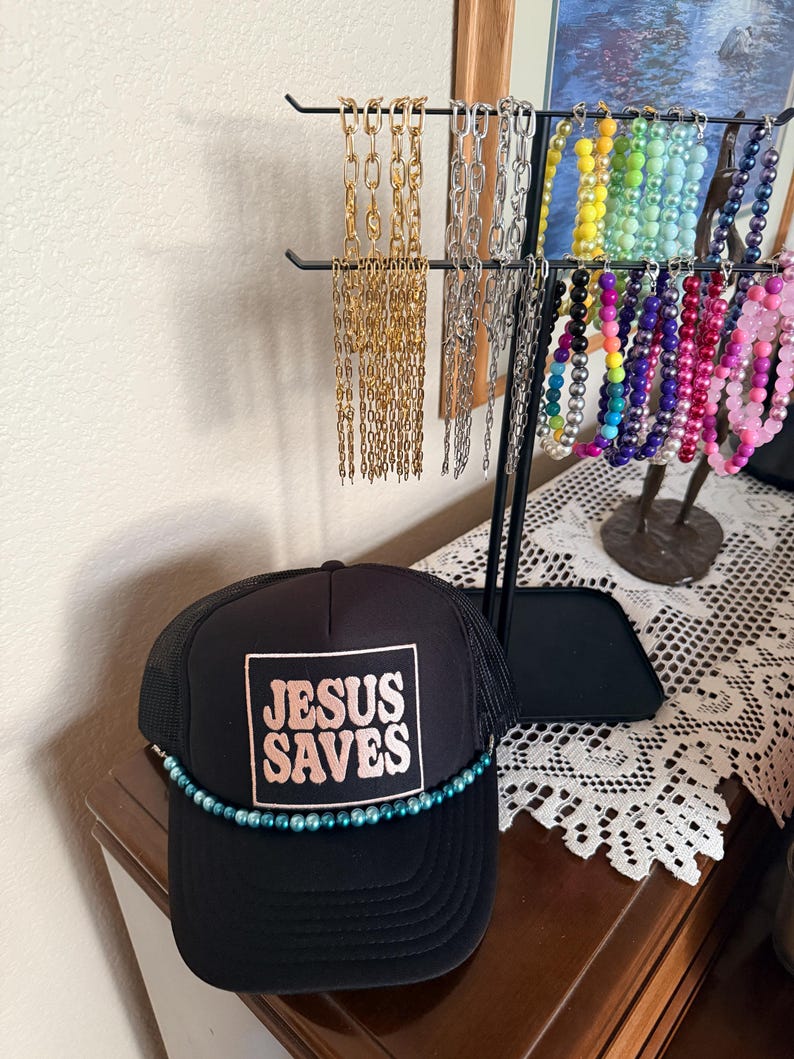 May include: Black trucker hat with the text "JESUS SAVES" in a square, with a teal bead trim. A jewelry display stand holds various necklaces in gold, silver, and colorful beads. The hat is on a wooden surface.