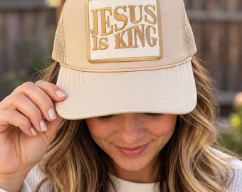 May include: A beige trucker hat featuring a rectangular patch with the text JESUS IS KING embroidered in gold thread. The cap includes a matching gold braided cord across the base of the crown and a mesh back panel for ventilation.