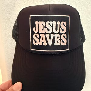 May include: Black trucker hat with a mesh back and a curved brim. The front features a square patch with the words "JESUS SAVES" in a pink, retro-style font.