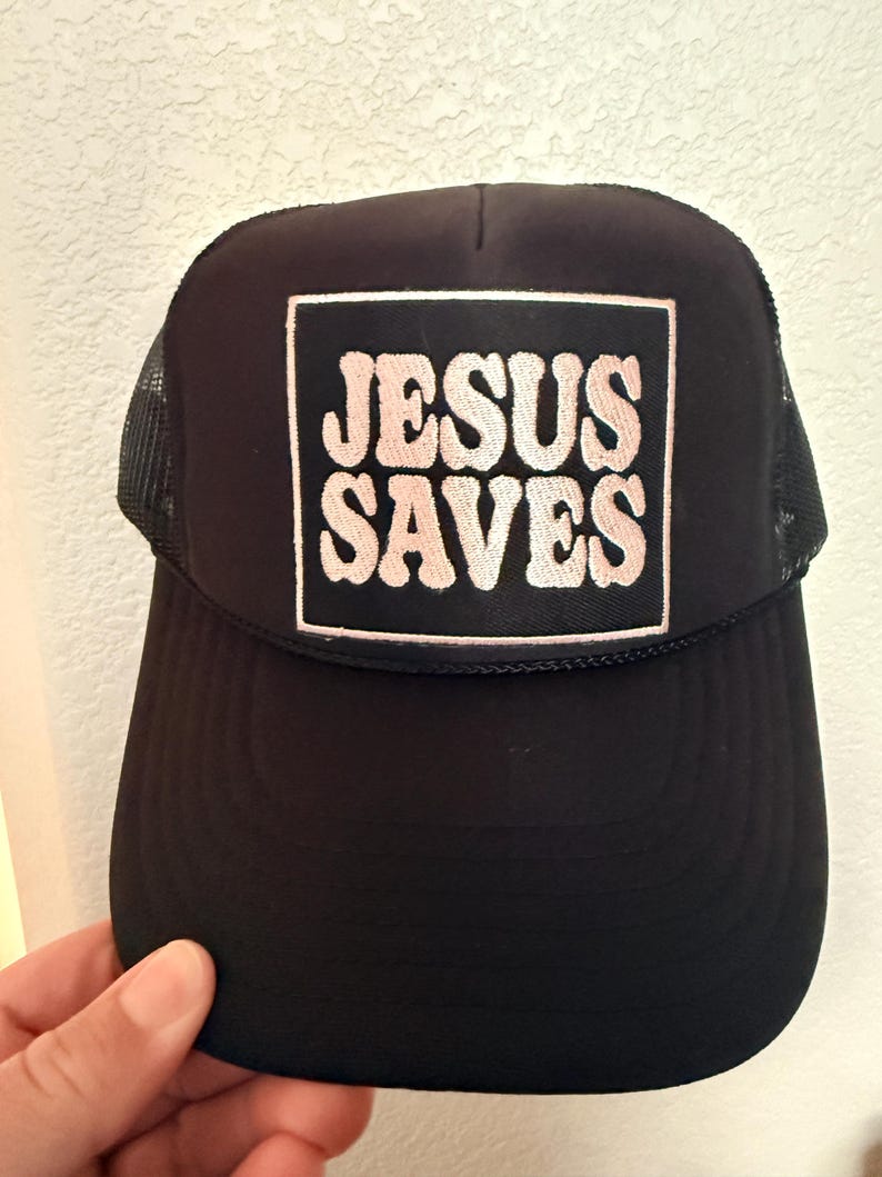 May include: Black trucker hat with a mesh back and a curved brim. The front features a square patch with the words "JESUS SAVES" in a pink, retro-style font.