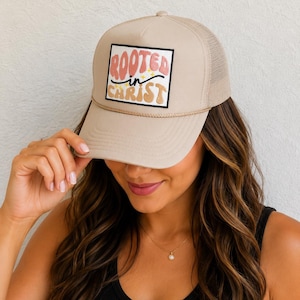 May include: Tan trucker hat with a mesh back and a curved brim. The front panel features a rectangular patch with the words "ROOTED IN CHRIST" in a colorful, retro font. A rope detail accents the brim.