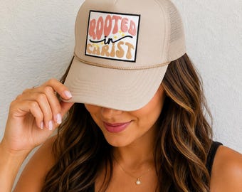 May include: Tan trucker hat with a mesh back and a curved brim. The front panel features a rectangular patch with the words "ROOTED IN CHRIST" in a colorful, retro font. A rope detail accents the brim.