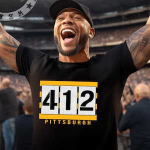 May include: Black t-shirt with the number "412" in white and gold, and the word "Pittsburgh" below. The shirt is worn by a person with arms raised in a stadium setting. The image includes a logo with crossed rifles.
