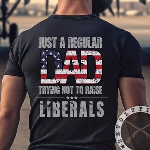May include: A black t-shirt with a distressed American flag design that says "Just a Regular Dad Trying Not to Raise Liberals". The shirt also has a small logo on the bottom right that says "Revolution Warfare".