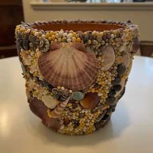 Może przedstawiać: Kwiatownik w kształcie kwadratu, ozdobiony mozaiką z muszli, małych kamyków i żółtych koralików. Doniczka posiada duże, brązowe muszle przegrzebków i naturalne, jasnobrązowe wnętrze. Zewnętrzna strona to mieszanka neutralnych tonów i faktur.