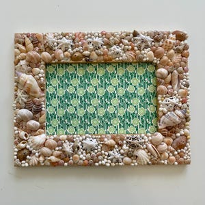 Puede incluir: Marco de fotos rectangular decorado con conchas marinas, estrellas de mar y pequeñas cuentas blancas. El interior del marco presenta un fondo con un patrón floral verde y blanco. Un artículo decorativo natural de temática costera.