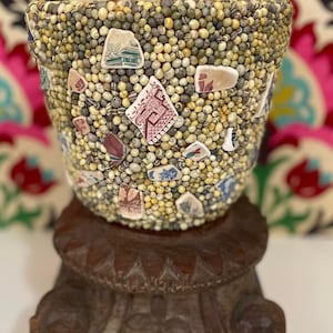 Puede incluir: Un cuenco de mosaico, elaborado con pequeñas cuentas redondas beige y verdes, adornado con fragmentos de baldosas de cerámica estampadas. El cuenco descansa sobre un soporte de madera tallada, mostrando una mezcla de texturas y colores.