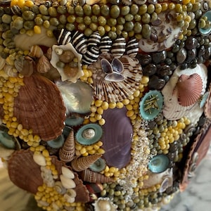 Puede incluir: Un cuenco decorativo cubierto con un mosaico de conchas marinas, pequeños guijarros y cuentas. El cuenco presenta una variedad de colores, incluyendo marrón, blanco, amarillo y púrpura. El diseño incluye conchas rayadas y pequeñas cuentas redondas.