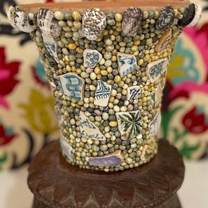 Puede incluir: Una maceta decorativa hecha a mano. El recipiente cilíndrico está cubierto de pequeñas piedras redondas y claras, adornado con conchas marinas y fragmentos de cerámica azul y blanca. Se apoya sobre un soporte de madera tallada, mostrando un diseño de mosaico único.