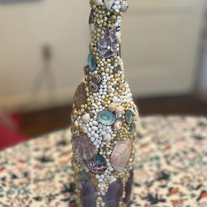 May include: A decorative bottle covered in various seashells, small white beads, and small tan beads. The bottle has a purple base and is adorned with a variety of shells in different shapes and sizes. The bottle is a unique piece of art.