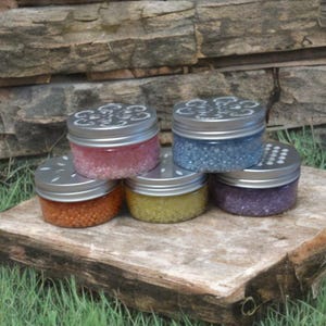 Könnte beinhalten: Fünf kleine Gläser mit silbernen Deckeln, die jeweils mit farbigem, körnigem Material gefüllt sind. Die Gläser sind auf einer Holzoberfläche vor einem Steinhintergrund angeordnet. Zu den Farben gehören Rosa, Blau, Orange, Gelb und Lila.