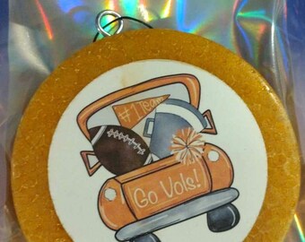 May include: A round, textured, golden-yellow air freshener in a clear plastic bag. The center features a graphic of an orange truck carrying a football, a megaphone, a pennant labeled #1 Team, and a pom-pom, with the text Go Vols! printed on the truck.