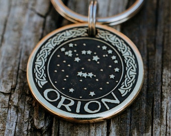 May include: A round, bronze-colored keychain pendant with the word "ORION" in white lettering. The pendant features a constellation design with stars and a Celtic knot border. A silver-colored key ring is attached.