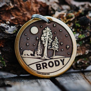 Op de afbeelding: Een hondenpenning van messing met een gegraveerd boslandschap. Het ontwerp bevat een beer, drie bomen, een maan en sterren. De penning is gegraveerd met de naam "Brody".