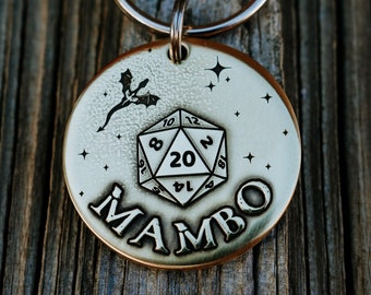 May include: A silver metal dog tag with a black and white illustration of a 20-sided die and a dragon. The tag is engraved with the word "MAMBO" in a stylized font.