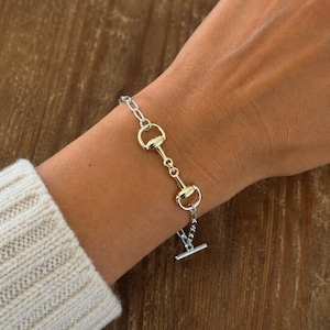 Op de afbeelding: Zilverkleurige armband met een paardenbit-ontwerp en goudkleurige accenten. De armband bestaat uit een ketting en een reeks bitvormige schakels. De T-bar sluiting is zichtbaar. De armband wordt om een pols gedragen.