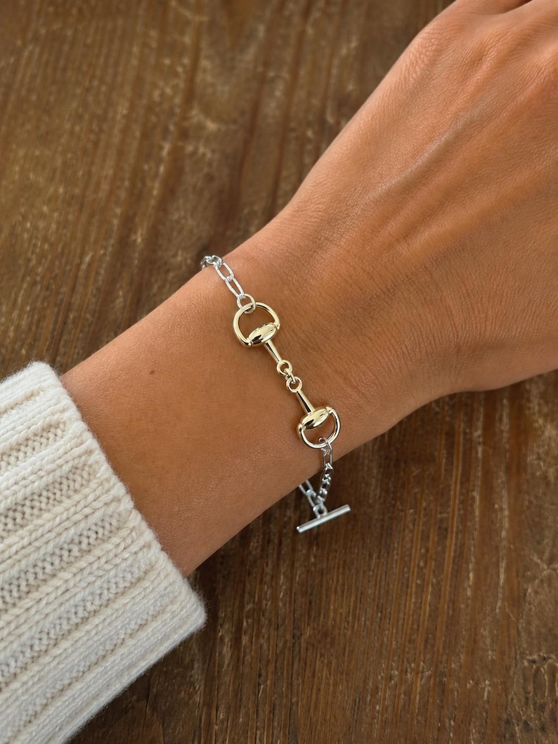 Op de afbeelding: Zilverkleurige armband met een paardenbit-ontwerp en goudkleurige accenten. De armband bestaat uit een ketting en een reeks bitvormige schakels. De T-bar sluiting is zichtbaar. De armband wordt om een pols gedragen.