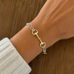 May include: A silver and gold-tone bracelet featuring a horse bit design. The bracelet is worn on a wrist, showcasing the contrasting colors and intricate details of the jewelry. The clasp is a T-bar style.