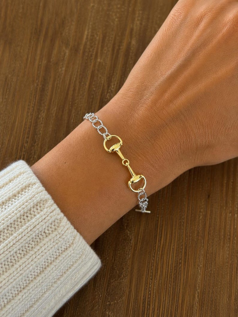 Op de afbeelding: Een tweekleurige armband met een paardenbit-ontwerp, gedragen om een pols. De armband combineert zilveren en goudkleurige elementen, met een T-bar sluiting. Het ontwerp doet denken aan paardensportaccessoires. De armband is ongeveer 18 cm lang.