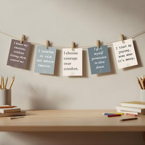 May include: A display of inspirational cards with positive affirmations, clipped to a string with wooden clothespins. The cards are in various colors, with phrases like "I honor my emotions" and "I choose courage over comfort."