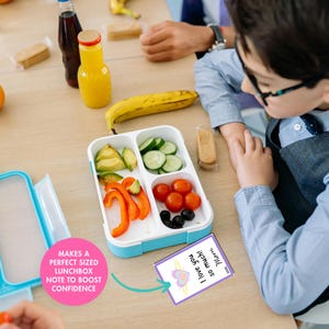 May include: A white and blue lunchbox filled with sliced vegetables, tomatoes, and olives. A note that says "I love you so much! Love, Mom" is next to the lunchbox. A banana and juice bottles are also on the table.