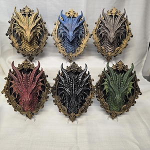 May include: A collection of six wall-mounted dragon head sculptures, each encased in an ornate, antique gold-colored oval frame. The dragon heads are rendered in distinct colors: gold, blue, grey, red, black, and green, featuring detailed scales and horns.