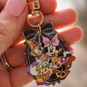 May include: A gold-toned metal keychain featuring a flat acrylic charm with a Halloween-themed illustration of Daisy Duck and Minnie Mouse. The characters are dressed in costumes, holding a jack-o'-lantern, framed by a black bow design.