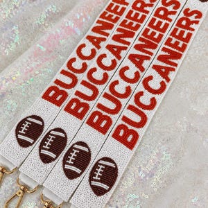 May include: A white beaded strap featuring the red text BUCCANEERS repeated vertically, interspersed with brown and white beaded football icons. The strap includes gold-toned metal swivel clasps at the bottom for attachment purposes.