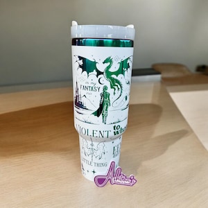 Puede incluir: Un vaso blanco con un diseño de fantasía verde y negro. El diseño incluye un dragón, una figura y el texto "in my fantasy era" y "Violent to what little thing". El vaso tiene una tapa plateada.