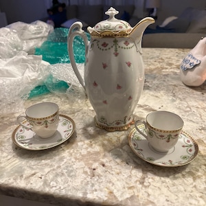 May include: A white porcelain teapot with a gold trim and floral design, along with two matching teacups and saucers.