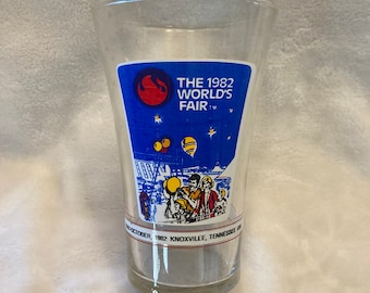 Peut inclure: Un verre en verre transparent avec un autocollant bleu et blanc représentant un logo de flamme rouge et le texte "THE 1982 WORLD'S FAIR TM". L'autocollant montre également une illustration de personnages de dessin animé dans une foire avec une montgolfière dans le ciel. Le texte "MAY - OCTOBER, 1982 KNOXVILLE, TENNESSEE USA" est imprimé au bas de l'autocollant.
