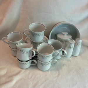 May include: A set of white porcelain teacups and saucers with a floral pattern. The set includes a sugar bowl with a lid and a creamer.