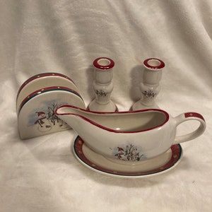 May include: A set of snowman themed ceramic kitchenware, including a gravy boat with a matching saucer, a napkin holder, and two candle holders. The pieces are decorated with a winter scene featuring snowmen and a red and green border.